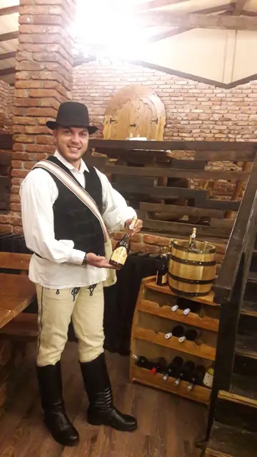 Ospătar în costum popular, lângă un raft cu vinuri